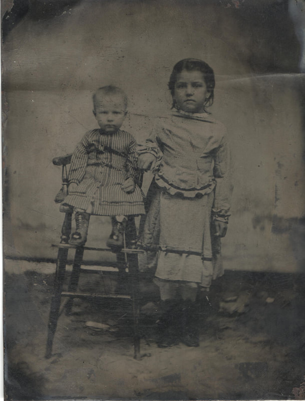 Baby in gown seated in high chair next to young girl standing