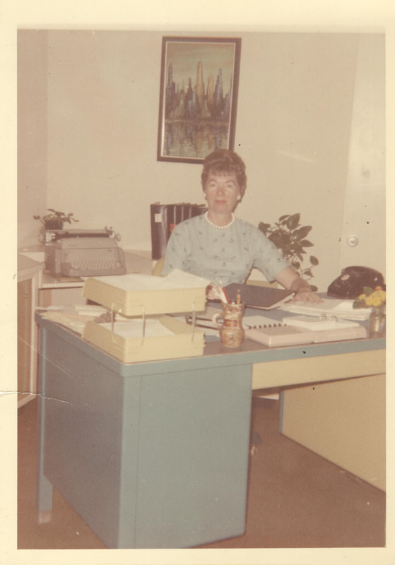 Woman in blue dress, necklace and earrings, seated a desk in office