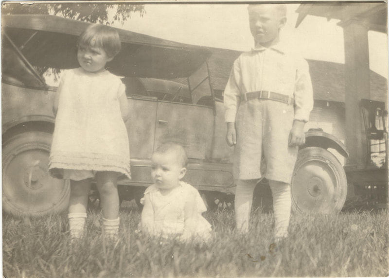 Baby seated between young boy and girl standing in front of car