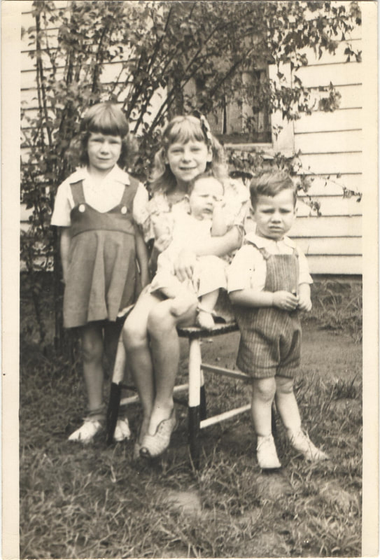 Small children in overalls standing next to girl holding young baby