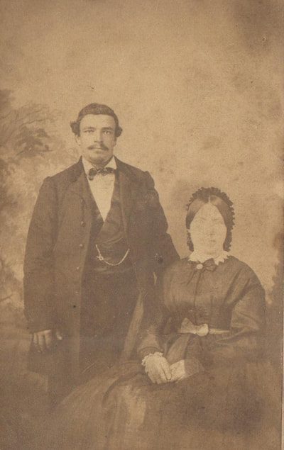 Man with mustache and three piece suit standing next to woman in full length dress seated